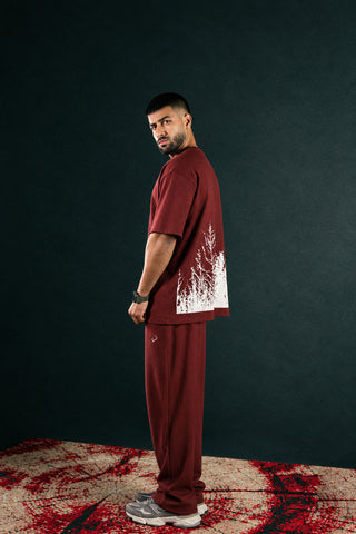 A man stands in profile wearing a matching rust-red t-shirt and sweatpants set with a white splatter graphic on the back of the shirt. The background is a dark textured wall with a red and black splatter-patterned floor.