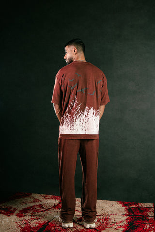 A man stands with his back to the camera wearing a brown t-shirt and matching brown sweatpants, featuring a white and dark graphic print on the back. He is standing on a patterned rug against a dark background.