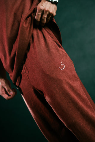 A close-up of a person wearing loose, draped rust-red pants and a matching top against a dark green background. A small white embroidered symbol is visible on the thigh.