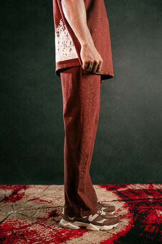 A person wearing a matching rust-brown short-sleeve t-shirt and wide-leg trousers, paired with brown and white sneakers, standing on a textured red and black rug against a dark background.