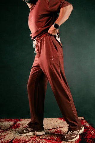 A person wearing a matching rust-brown lounge set consisting of a short-sleeve t-shirt and drawstring sweatpants, standing on a patterned rug against a dark green background.