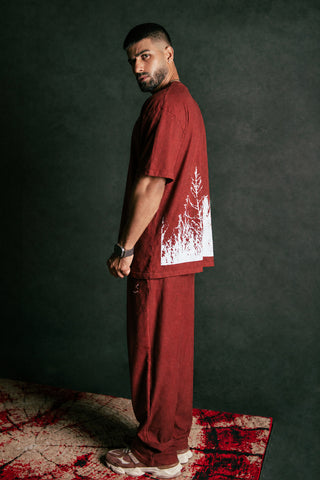 A man stands in profile wearing a matching rust-red t-shirt and pants set with a white splatter graphic on the shirt, set against a dark textured studio background.