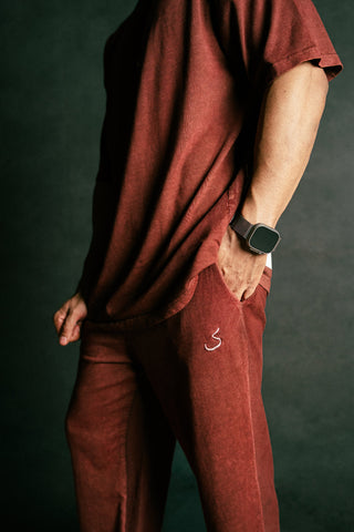 A person wearing a matching rust-red short-sleeve top and jogger pants set, with the pants featuring a small white embroidered logo on the left thigh. The person is shown from the chest down against a dark textured background, wearing a black smartwatch.