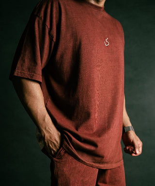 Wearing an oversized rust-red short-sleeve t-shirt and matching pants, standing against a dark studio background. The shirt has a small white stylized logo on the left chest.