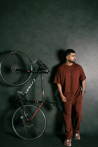 A man wearing a matching rust-brown t-shirt and sweatpants stands next to a black road bicycle with red accents against a dark textured studio background.