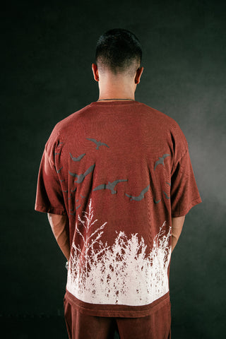 A man viewed from behind wearing a rust-brown t-shirt with a white grass-like graphic and dark bird silhouettes on the back, paired with matching brown pants, set against a dark studio background.
