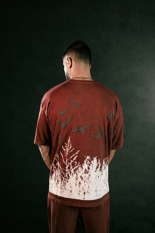 A man viewed from behind wearing a rust-red t-shirt with a white forest and bird graphic on the back, set against a dark textured background.