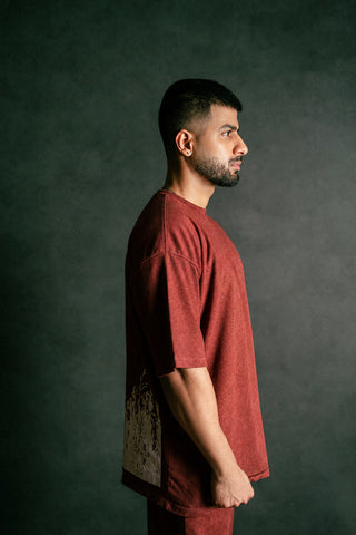 A man in profile wearing a rust-red oversized t-shirt and matching pants, standing against a dark textured studio background.