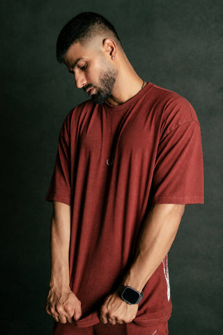 A man wearing a matching rust red t-shirt and sweatpants set, looking down against a dark textured studio background.